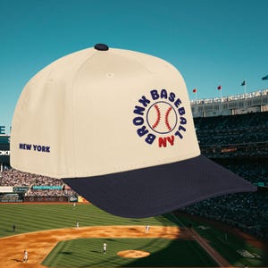 May include: Cream-colored baseball cap with a navy blue brim and top button. The cap features the text "BRONX BASEBALL NY" in a circular design with a baseball graphic. The words "NEW YORK" are printed on the side.