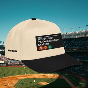 May include: A cream-colored baseball cap with a black brim. The cap features the text "161 Street-Yankee Stadium Station" and the letters "B D 4". The text "NEW YORK" is also visible on the side.