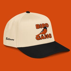 Może przedstawiać: Kremowa czapka bejsbolowa z czarnym daszkiem. Czapka posiada haftowaną pomarańczową grafikę ptaka oraz napisy "BIRD GANG". Z boku wyhaftowano słowo "Baltimore".