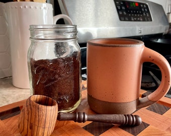 May include: A wooden scoop with a brown and black wood grain pattern sits on a checkered cutting board next to a glass jar filled with coffee grounds and a speckled light brown ceramic mug.