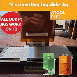May include: A dog tag slider jig with the text "19 x 3 mm Dog Tag Slider Jig" and "ALL OUR F1 JIGS WORK ON F2". The image shows a laser engraving machine with a gray tray and several colored jigs. Two small XTOOL machines are also visible.