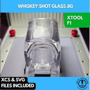 May include: A clear glass shot glass jig for laser engraving. The jig is made of metal and has a square base. The jig is designed to hold a shot glass in place for engraving. The image includes the text "XTOOL F1" and "XCS & SVG FILES INCLUDED".