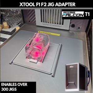 Op de afbeelding: XTOOL F1 F2 Jig Adapter met een helder glas en roze laserlicht. De tekst "XTOOL F1 F2 JIG ADAPTER" staat bovenaan. De tekst "ENABLES OVER 300 JIGS" staat onderaan. De afbeelding bevat ook een CREALITY FALCON T1 lasergraveerder.