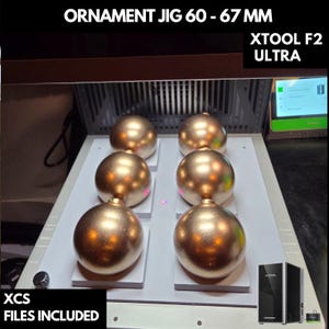 May include: Six gold-colored Christmas ornaments, each approximately 2.5 inches in diameter, are arranged on a white platform. The ornaments are part of an ornament jig, with the text "ORNAMENT JIG 60 - 67 MM" and "XCS FILES INCLUDED" visible.
