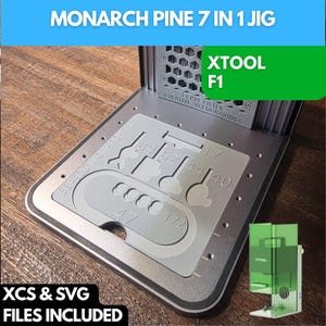 May include: A silver metal jig with the text "MONARCH PINE 7 IN 1 JIG" and "XTOOL F1". The jig includes various cutouts and numbers, with the words "XCS & SVG FILES INCLUDED". A small green and clear XTOOL F1 is also visible.