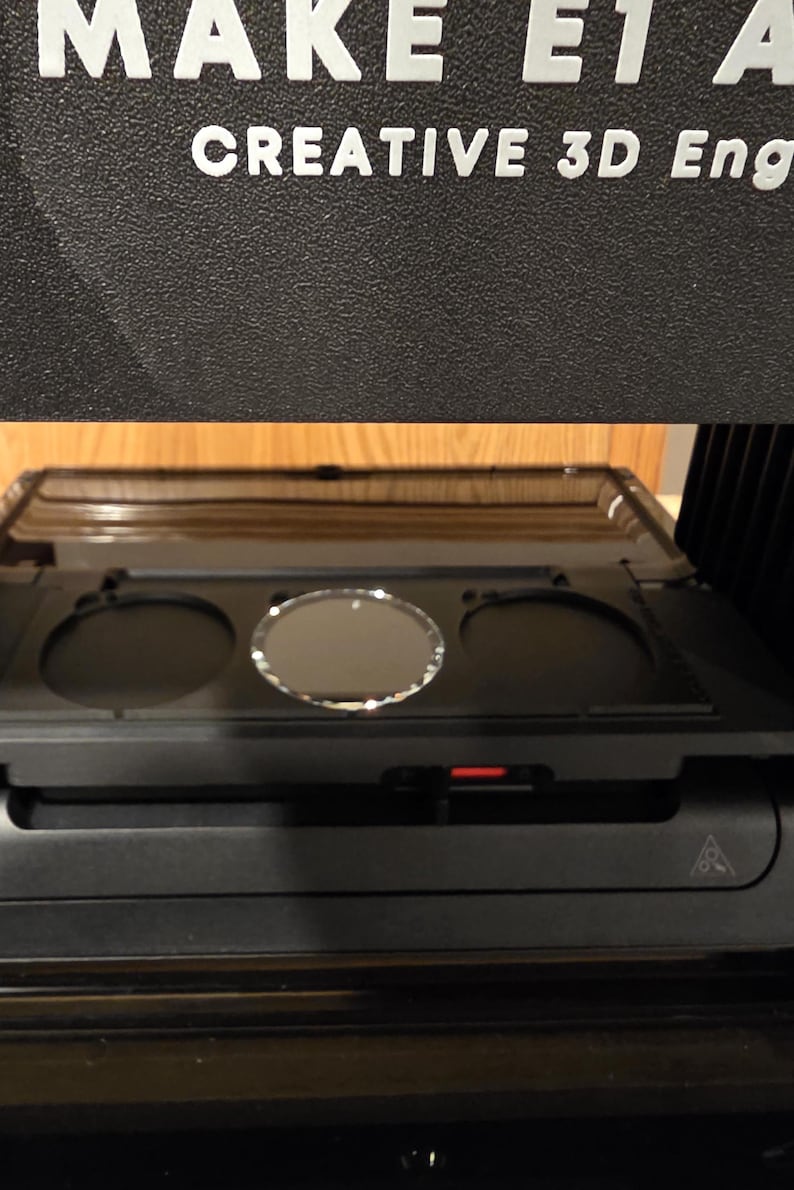 May include: A black 3D printer with the text "MAKE E1 A" and "CREATIVE 3D Eng" in white. The printer has a tray with three circular openings and a red indicator light.
