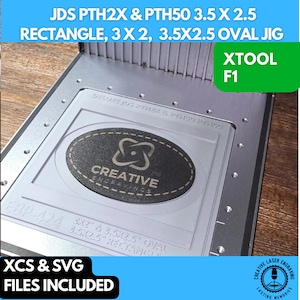 May include: A silver metal jig with two cutouts for laser engraving. The larger cutout is oval shaped and measures 3.5 inches by 2.5 inches. The smaller cutout is rectangular and measures 3 inches by 2 inches. The jig is labeled "JDS PTH2X & PTH50 3.5 X 2.5 RECTANGLE, 3 X 2, 3.5X2.5 OVAL JIG" and "XTOOL F1".