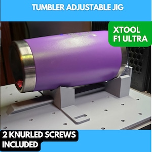 May include: A purple tumbler sits in a 3D printed adjustable jig with two knurled screws included. The jig is designed to hold the tumbler in place for engraving or other customization. The text "XTOOL F1 ULTRA" is visible in the upper right corner of the image.