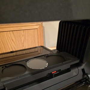 May include: A black countertop appliance with a clear glass lid and three circular openings. A clear, oval-shaped object sits in the center opening. The appliance has a red power indicator light.
