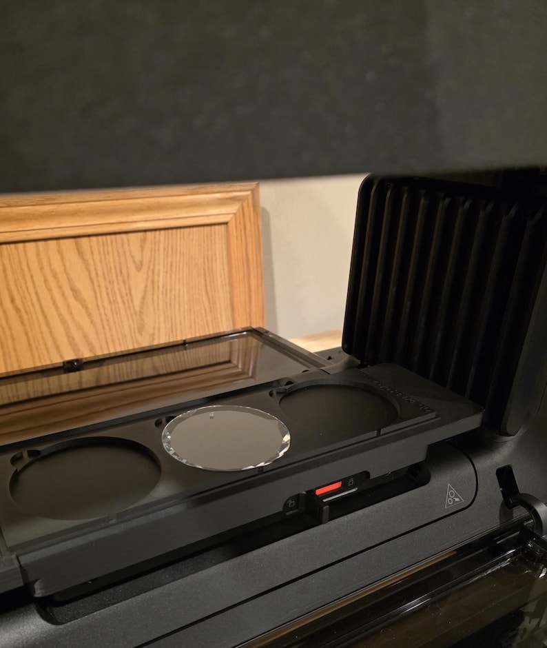 May include: A black countertop appliance with a clear glass lid and three circular openings. A clear, oval-shaped object sits in the center opening. The appliance has a red power indicator light.