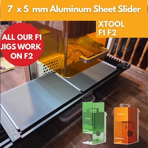 May include: A silver and black XTOOL laser cutter with a 7 x 5 mm aluminum sheet slider. The image includes the text "ALL OUR F1 JIGS WORK ON F2" and "XTOOL F1 F2". Two smaller, transparent laser cutter models are also shown.
