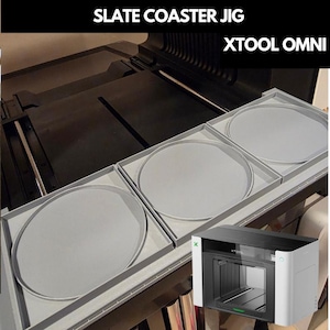 May include: A grey slate coaster jig for the xTool Omni laser machine, featuring three square trays with circular indentations for holding coasters. The text SLATE COASTER JIG and XTOOL OMNI appears at the top, with a small xTool machine image.