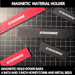 Puede incluir: Soportes magnéticos para materiales en rojo y negro, con el texto "STAYDOWNZ" en blanco. La imagen muestra varias barras rectangulares, algunas de 15 cm y 7.6 cm de largo, diseñadas para camas de panal y metal.