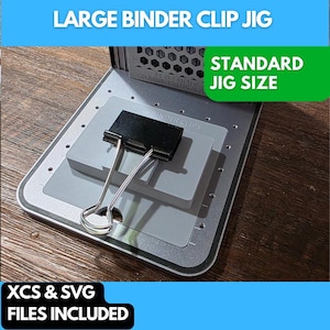 May include: A silver and gray jig with a black binder clip. The text "LARGE BINDER CLIP JIG" is at the top, with "STANDARD JIG SIZE" in a green box. The text "XCS & SVG FILES INCLUDED" is at the bottom.