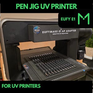Peut inclure: Une imprimante UV noire et argentée avec un accessoire de jig pour stylo. Le jig maintient plusieurs stylos en place pour la gravure. L'imprimante possède un écran d'affichage noir et blanc et une étiquette noire et blanche qui indique "EUFYMAKE E1 AP ADAPTER CREATIVE 3D Engravings".