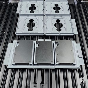 May include: Three black metal wallets with the text "SKAGEN DENMARK" and "012810" on them are displayed in a gray plastic holder. The holder has four slots, and the wallets are in the first three slots.