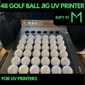 Op de afbeelding: Een zwarte UV-printer met een mal voor 48 golfballen. De mal is gevuld met witte golfballen. De printer heeft een label met de tekst "EUFY E1 M".