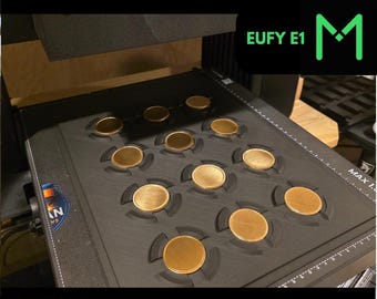 May include: A black coin jig with multiple circular slots holding gold-colored coins. The text "40 - 50 MM COIN JIG" and "FOR UV PRINTERS" are displayed in green. The jig is designed for coins approximately 1.57 to 1.97 inches in diameter.