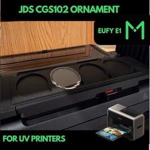 May include: A black UV printer with a tray displaying a printed image. The image also shows a clear, round ornament and the text "JDS CGS102 ORNAMENT" and "FOR UV PRINTERS". The printer is next to a black tray with circular cutouts.