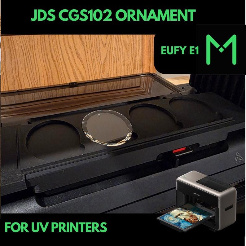 May include: A black UV printer with a tray displaying a printed image. The image also shows a clear, round ornament and the text "JDS CGS102 ORNAMENT" and "FOR UV PRINTERS". The printer is next to a black tray with circular cutouts.