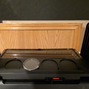 May include: A black audio component with a clear, oval-shaped glass disc in one of the circular recesses. The component has a rectangular tray with three circular indentations. The background features a wooden cabinet.