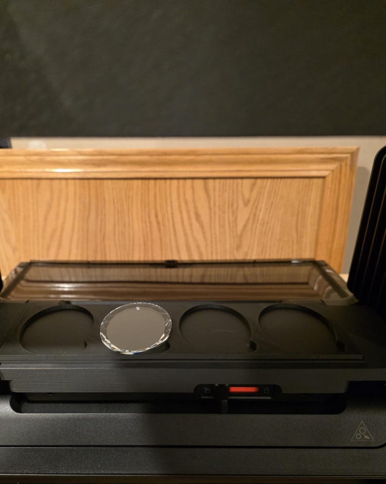 May include: A black audio component with a clear, oval-shaped glass disc in one of the circular recesses. The component has a rectangular tray with three circular indentations. The background features a wooden cabinet.