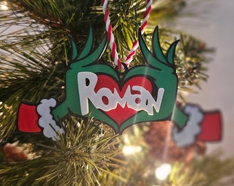 Puede incluir: Un adorno navideño verde y rojo con el nombre "Roman" escrito en letras blancas dentro de un corazón rojo. El adorno tiene forma de dos manos sosteniendo el corazón.
