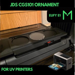 May include: A black UV printer with the text "JDS CGS101 ORNAMENT" and "FOR UV PRINTERS" in green. The printer has a clear top and a small silver and black printer on the side with a printed image.
