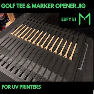 May include: A black tray with multiple slots holds a row of beige golf tees. The text "GOLF TEE & MARKER OPENER JIG" and "FOR UV PRINTERS" are displayed in green. The logo "M" is also visible.