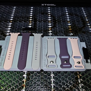May include: Five different colored watch bands for a smartwatch. The bands are in a light gray holder with a white label that says "XTOOL". The bands are pink, purple, gray, purple, and pink.