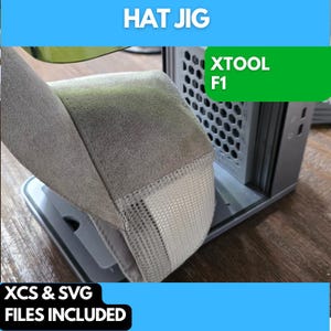 May include: A gray and white 3D printed hat jig for a computer case. The jig is designed to hold a hat in place while it is being printed. The jig is labeled "XTOOL F1".