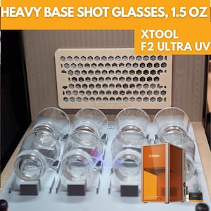 May include: Clear heavy base shot glasses, 1.5 oz capacity, arranged in a row. The glasses are displayed with an XTOOL F2 ULTRA UV laser engraving machine. The text "HEAVY BASE SHOT GLASSES, 1.5 OZ" is visible at the top.