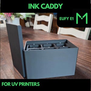 Peut inclure: Un bac à encre gris foncé avec le texte "INK CADDY" et "FOR UV PRINTERS" en vert. Le bac comporte plusieurs compartiments étiquetés avec des lettres et un couvercle à charnière. L'image comprend également un logo avec le texte "EUFY E1" et un "M" vert.