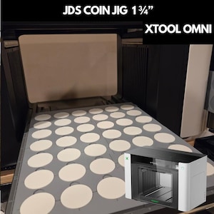 Op de afbeelding: Een grijze JDS Coin Jig 1 3/4" voor de xTool Omni lasermachine, voorzien van een raster met ronde uitsparingen voor het vasthouden van munten. De mal is geplaatst in de zwart-witte xTool Omni lasermachine met een glazen deur en verlichting.