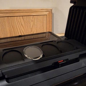 May include: A black panini press with a clear glass lid and a circular glass object inside. The press is open, revealing the cooking surface and a red power indicator. The background includes a wooden cabinet.
