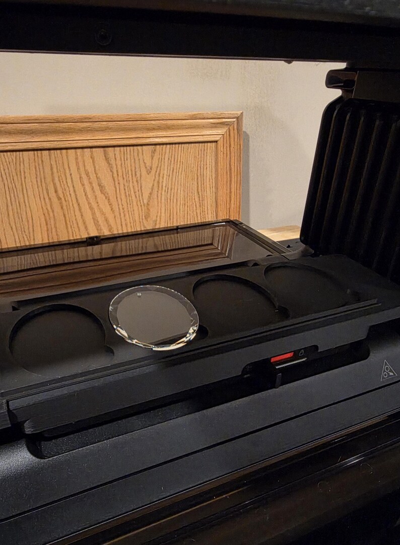 May include: A black panini press with a clear glass lid and a circular glass object inside. The press is open, revealing the cooking surface and a red power indicator. The background includes a wooden cabinet.