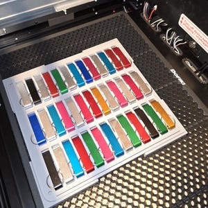 May include: A tray of 36 colorful metal keychains, each with a different color and finish. The keychains are arranged in rows and columns on a white tray. The tray is sitting on a black surface with a honeycomb pattern.