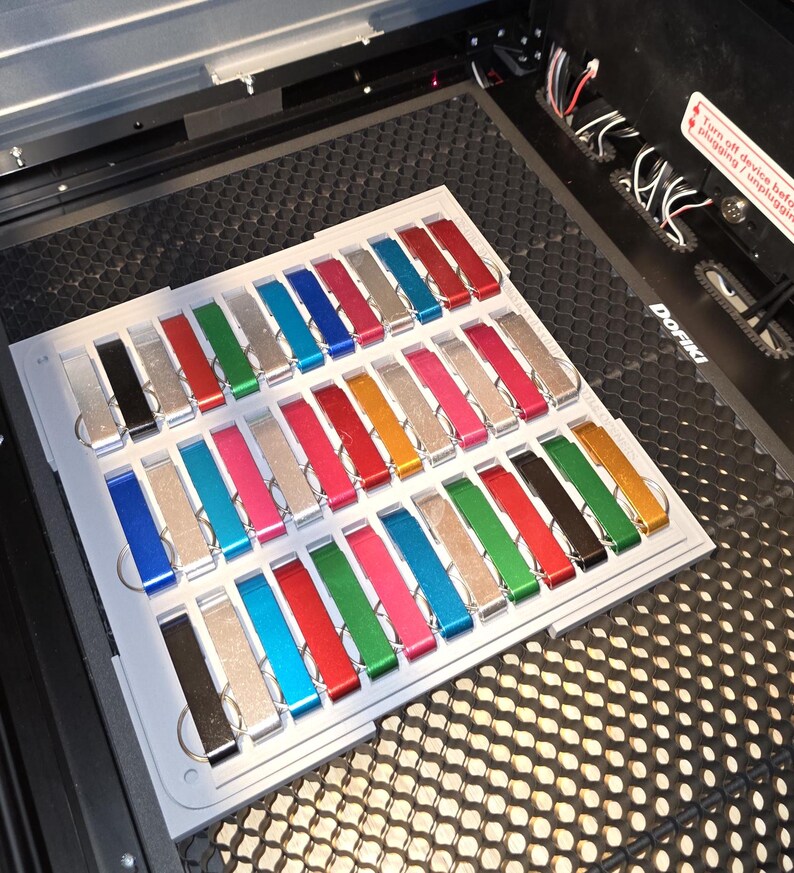 May include: A tray of 36 colorful metal keychains, each with a different color and finish. The keychains are arranged in rows and columns on a white tray. The tray is sitting on a black surface with a honeycomb pattern.