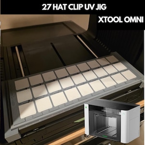Op de afbeelding: Een 27-delige hoedclip UV-mal voor de XTool Omni, met een rechthoekig grijs rooster met zevenentwintig witte vierkante inzetstukken. De tekst 27 HAT CLIP UV JIG en XTOOL OMNI staat bovenaan. Een kleine inzet toont de witte en zwarte XTool machine.