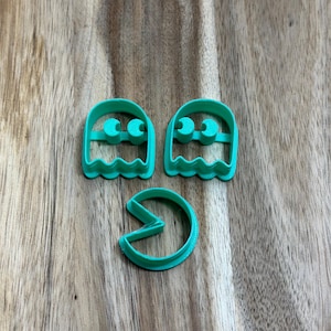 May include: Three green plastic cookie cutters shaped like Pac-Man and two ghosts. The ghosts have two black dots for eyes.