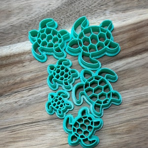 May include: Set of six green plastic turtle cookie cutters of varying sizes. The cutters are arranged in a cluster on a wooden surface.