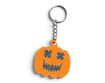 Puede incluir: Un llavero de calabaza naranja brillante con purpurina que presenta una cara de jack-o'-lantern con dos ojos negros brillantes en forma de X y una boca negra dentada. La calabaza está unida a una anilla de metal plateado mediante una cadena.