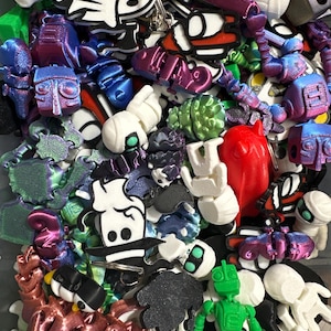 May include: A collection of colorful 3D printed keychains featuring various designs, including robots, animals, and abstract shapes. The keychains are made of different materials, including plastic and metal.