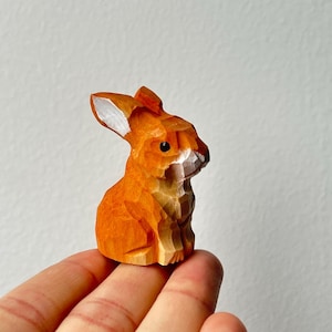 Op de afbeelding: Een kleine, handgesneden houten konijnenfiguur. Het konijn heeft een warme oranje kleur met gedetailleerde gravures, die zijn oren, gezicht en lichaam laten zien. Het beeldje wordt in een hand gehouden, wat de grootte en het vakmanschap benadrukt.