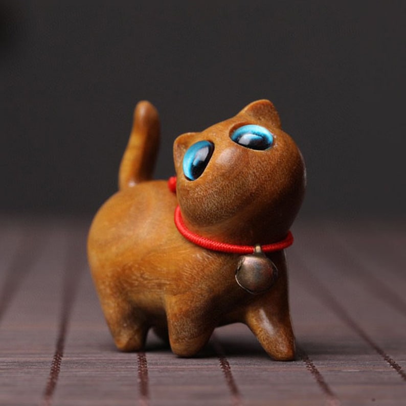 Peut inclure: Une figurine de chat en bois avec de grands yeux bleus et un collier rouge avec une clochette. Le chat est debout sur une surface brune.