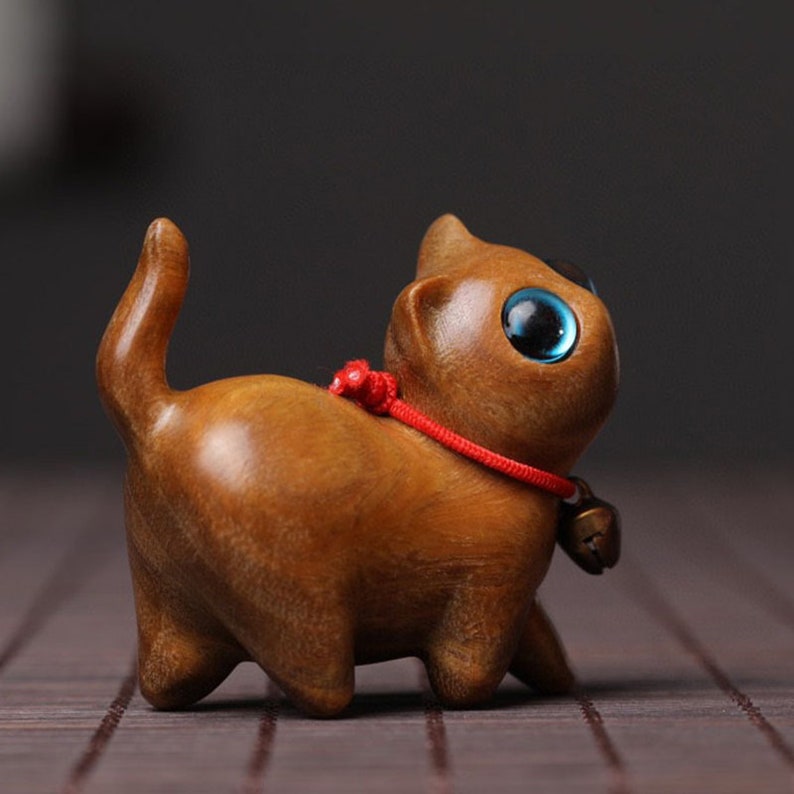 Peut inclure: Une petite figurine de chat en bois avec une ficelle rouge attach&eacute;e autour du cou et une cloche suspendue &agrave; la ficelle. Le chat a de grands yeux bleus et un corps en bois brun.