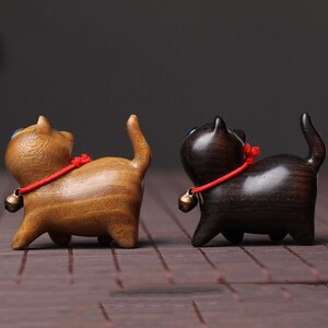 Peut inclure: Deux figurines de chat en bois, l'une brune et l'autre noire, avec des colliers en ficelle rouge et des cloches. Les chats sont tourn&eacute;s dans des directions diff&eacute;rentes et ont une expression espi&egrave;gle.