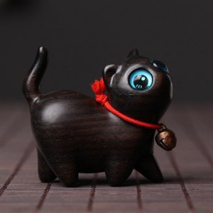 Peut inclure: Une petite figurine de chat en bois noir avec des yeux bleus et un collier rouge en ficelle avec une clochette. La figurine est debout sur une surface brune.