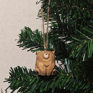 Könnte beinhalten: Ein hölzerner Bären-Weihnachtsbaumschmuck mit natürlichem Finish, der an einer Schnur hängt. Der Bär ist mit geometrischen Formen geschnitzt und hat eine weiße Nase und kleine schwarze Pfoten. Der Schmuck hängt an einem grünen Weihnachtsbaum.