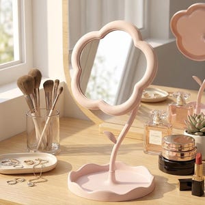 Può includere: Uno specchio da toeletta rosa a forma di fiore con un supporto a forma di stelo e una piccola base a vassoio. Pennelli per il trucco, gioielli e flaconi di profumo sono disposti su una superficie di legno.